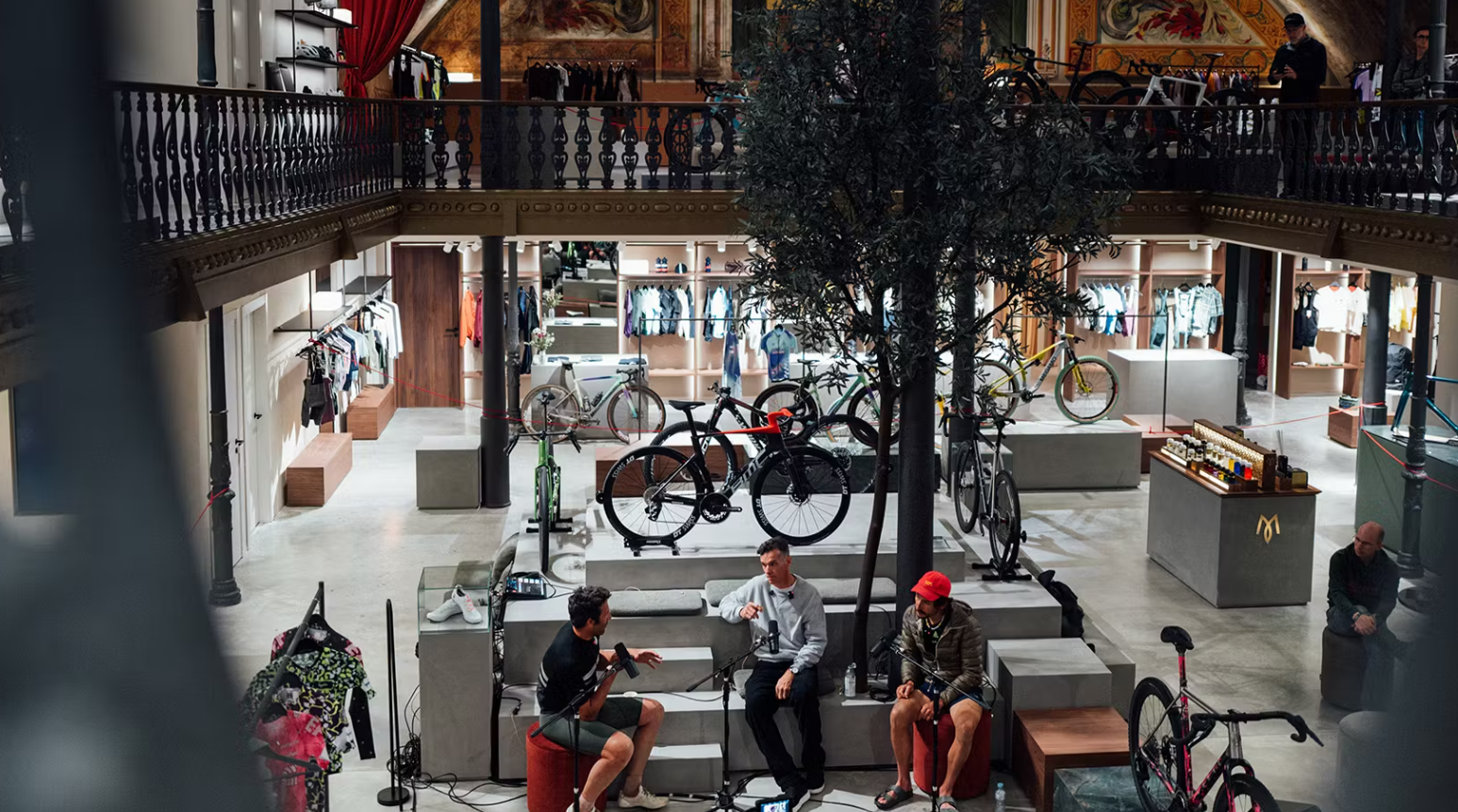 a modern bike shop with bicycles, cycling gear, and apparel displays. The space features a mezzanine level, elegant railings, and a large indoor tree adding a stylish touch.